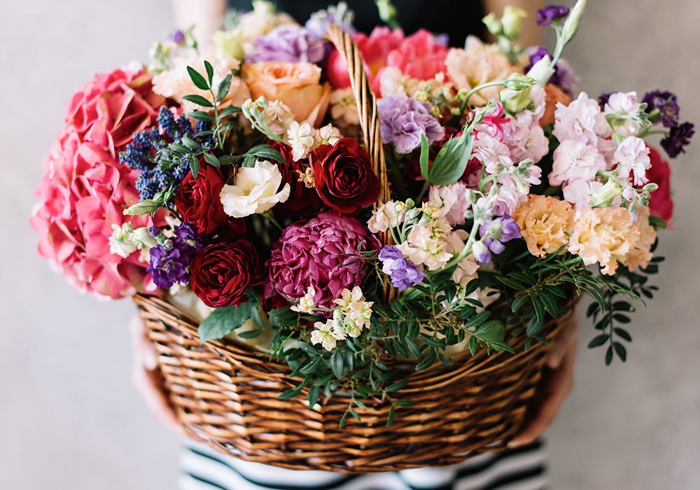 A large wicker basket containing an assortment of colorful flowers, including roses and hydrangeas, against a neutral background.